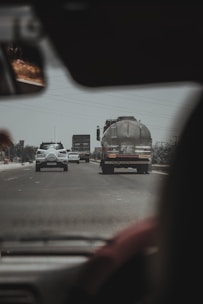 A view from inside a vehicle looking out at a road with traffic. In the foreground, there's part of the interior of the vehicle, including a rearview mirror. On the road, several vehicles are visible, including a tanker truck with text on the back and a few cars moving in each direction. Power lines run overhead and there's an overcast sky.