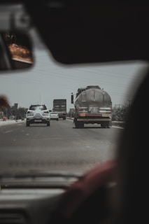 A view from inside a vehicle looking out at a road with traffic. In the foreground, there's part of the interior of the vehicle, including a rearview mirror. On the road, several vehicles are visible, including a tanker truck with text on the back and a few cars moving in each direction. Power lines run overhead and there's an overcast sky.