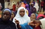 A group of women dressed in colorful veils and robes are gathered together. Some are interacting with each other, while others are focused on something in the distance. The background shows more people, potentially in a social or communal setting.
