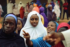 A group of women dressed in colorful veils and robes are gathered together. Some are interacting with each other, while others are focused on something in the distance. The background shows more people, potentially in a social or communal setting.