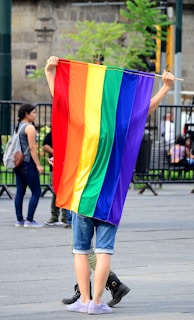 A smiling person holding a rainbow flag, standing in a peaceful park setting.