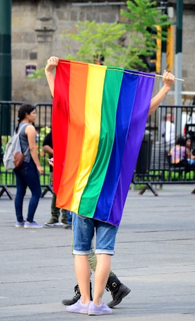 A smiling person holding a rainbow flag, standing in a peaceful park setting.