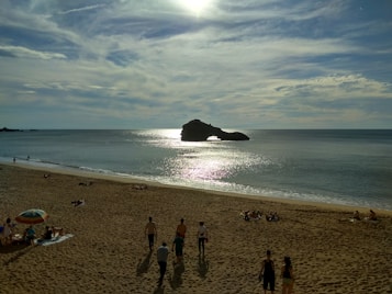 The image features a sandy beach with several people relaxing and walking. A distinctive rock formation is visible on the horizon, silhouetted against the shimmering water as the sun reflects off the surface. The sky is mostly clear with some clouds, creating a serene and expansive backdrop.