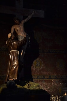 A statue depicting a robed figure reaching towards Jesus on the cross, set against a richly decorated background. The scene is dimly lit, highlighting the somber atmosphere.