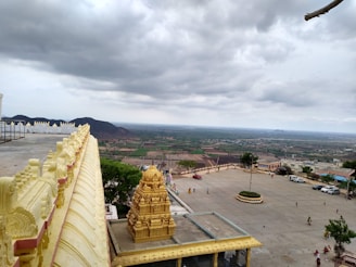 A temple with richly decorated golden architectural features is seen in the foreground, overlooking a vast open landscape of fields and distant hills under an overcast sky. There is a parking area with several vehicles and a few people walking around. The expansive view and prominent temple structure create a sense of serenity and spirituality.