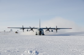A large military aircraft is landing on a snowy and icy terrain creating a cloud of snow and mist around its rear. The wide wingspan with multiple propellers is clearly visible. Various structures, possibly research facilities, are in the background along with a large radar dome. The horizon is flat, with a clear blue sky and no visible signs of vegetation.