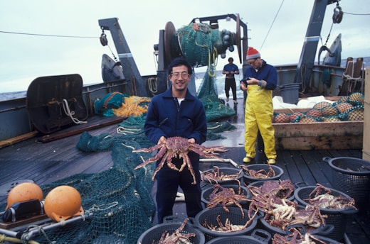 A person holding a large crab stands on the deck of a fishing boat. Surrounding them are baskets filled with crabs and fishing equipment, including nets and buoys. Another person in yellow waterproof overalls works in the background near a pile of nets.