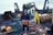 A person holding a large crab stands on the deck of a fishing boat. Surrounding them are baskets filled with crabs and fishing equipment, including nets and buoys. Another person in yellow waterproof overalls works in the background near a pile of nets.
