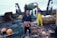 A person holding a large crab stands on the deck of a fishing boat. Surrounding them are baskets filled with crabs and fishing equipment, including nets and buoys. Another person in yellow waterproof overalls works in the background near a pile of nets.