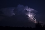 A dark night sky is illuminated by a bright bolt of lightning striking through large, ominous storm clouds. The scene is dramatic and powerful, with the silhouette of power lines intersecting the view. The landscape below is darkened, with the outline of trees barely visible.