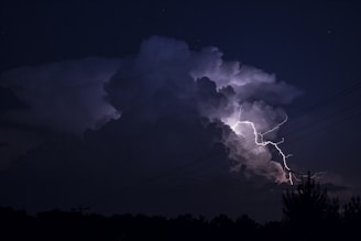 A dark night sky is illuminated by a bright bolt of lightning striking through large, ominous storm clouds. The scene is dramatic and powerful, with the silhouette of power lines intersecting the view. The landscape below is darkened, with the outline of trees barely visible.