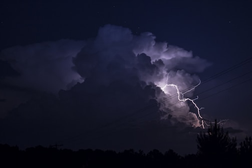 A dark night sky is illuminated by a bright bolt of lightning striking through large, ominous storm clouds. The scene is dramatic and powerful, with the silhouette of power lines intersecting the view. The landscape below is darkened, with the outline of trees barely visible.