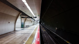 A deserted subway station platform with clean, tiled walls and a brightly lit ceiling. The platform is empty, and the tracks lead into a dark tunnel. Overhead signs and safety markings are visible, adding to the sense of order and functionality.
