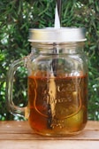 A glass mason jar with a handle is filled with a translucent amber liquid. It is topped with a silver metal lid and has a straw inserted through the lid. The jar is set against a leafy green background and rests on a wooden surface.