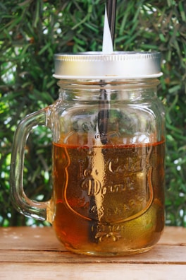 A glass mason jar with a handle is filled with a translucent amber liquid. It is topped with a silver metal lid and has a straw inserted through the lid. The jar is set against a leafy green background and rests on a wooden surface.