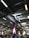 A large indoor space with an industrial-style ceiling featuring exposed ductwork and fluorescent lighting. Below, there are various goods and displays typical of a hardware or retail store with signs and product displays visible. A prominent purple column stands in the middle, surrounded by different types of merchandise.