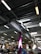 A large indoor space with an industrial-style ceiling featuring exposed ductwork and fluorescent lighting. Below, there are various goods and displays typical of a hardware or retail store with signs and product displays visible. A prominent purple column stands in the middle, surrounded by different types of merchandise.