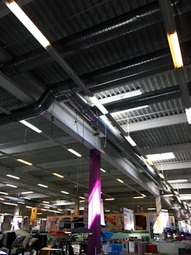 A large indoor space with an industrial-style ceiling featuring exposed ductwork and fluorescent lighting. Below, there are various goods and displays typical of a hardware or retail store with signs and product displays visible. A prominent purple column stands in the middle, surrounded by different types of merchandise.