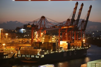 A cargo ship is docked at a port during twilight, with several large cranes towering above it. The ship is loaded with numerous shipping containers, and the scene is illuminated by artificial lights, casting a warm glow over the area. The sky shows the transition from day to night with a darkening horizon.