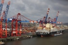 A large cargo ship loaded with colorful shipping containers is docked at a busy industrial port. Several towering cranes in blue and red are actively engaged in loading or unloading the containers onto the ship. The port is filled with stacks of containers, showcasing a bustling hub of maritime trade.