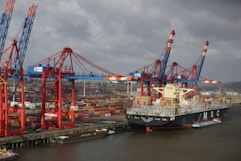 A large cargo ship loaded with colorful shipping containers is docked at a busy industrial port. Several towering cranes in blue and red are actively engaged in loading or unloading the containers onto the ship. The port is filled with stacks of containers, showcasing a bustling hub of maritime trade.