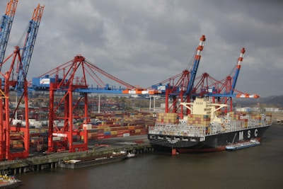 A large cargo ship loaded with colorful shipping containers is docked at a busy industrial port. Several towering cranes in blue and red are actively engaged in loading or unloading the containers onto the ship. The port is filled with stacks of containers, showcasing a bustling hub of maritime trade.