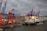 A large cargo ship loaded with colorful shipping containers is docked at a busy industrial port. Several towering cranes in blue and red are actively engaged in loading or unloading the containers onto the ship. The port is filled with stacks of containers, showcasing a bustling hub of maritime trade.