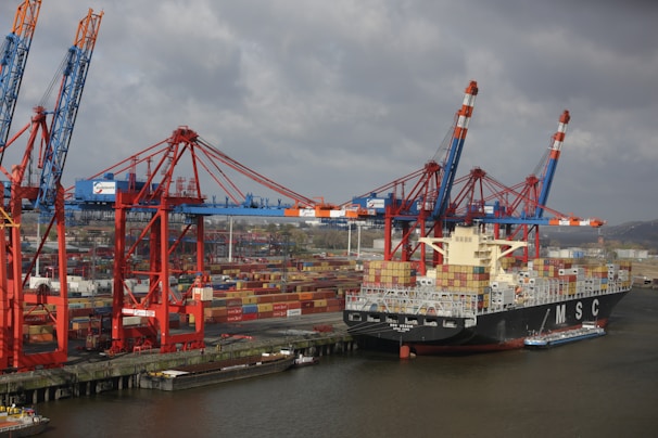 A large cargo ship loaded with colorful shipping containers is docked at a busy industrial port. Several towering cranes in blue and red are actively engaged in loading or unloading the containers onto the ship. The port is filled with stacks of containers, showcasing a bustling hub of maritime trade.