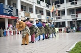 A group of people in colorful traditional clothing are standing in a school courtyard, each with a sarong wrapped around their waist. They are leaning on each other in a coordinated manner, creating a line. The background shows a multi-story school building with students and staff observing the performance. Flags are hoisted on poles in the background.