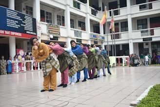 A group of people in colorful traditional clothing are standing in a school courtyard, each with a sarong wrapped around their waist. They are leaning on each other in a coordinated manner, creating a line. The background shows a multi-story school building with students and staff observing the performance. Flags are hoisted on poles in the background.