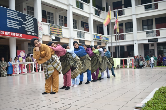 A group of people in colorful traditional clothing are standing in a school courtyard, each with a sarong wrapped around their waist. They are leaning on each other in a coordinated manner, creating a line. The background shows a multi-story school building with students and staff observing the performance. Flags are hoisted on poles in the background.