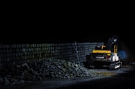 A construction vehicle is parked next to a stack of rocks and a stone wall, illuminated against a dark background. The vehicle appears to be a yellow and black excavator, partially surrounded by scattered stones.