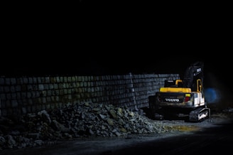 A construction vehicle is parked next to a stack of rocks and a stone wall, illuminated against a dark background. The vehicle appears to be a yellow and black excavator, partially surrounded by scattered stones.