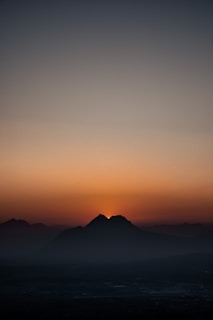 A serene sunrise with the sun emerging behind a mountain range, casting a soft orange and red glow across the sky. The mountains are silhouetted against the vibrant sky, and subtle mist envelops the lower areas, giving a tranquil and majestic view.