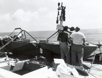 Several individuals are working on a boat or small vessel on a wooden deck, overlooking the ocean. The atmosphere suggests a maritime setting with the presence of equipment, possibly for survey or research. The sign indicates a survey buoy, suggesting a scientific or technical purpose.