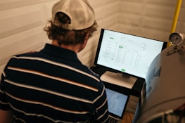 A person with a striped shirt and a cap is standing in front of a computer screen displaying data and analytics. Another laptop is open on a desk below the monitor, and a large metallic tank with a pressure gauge is nearby.