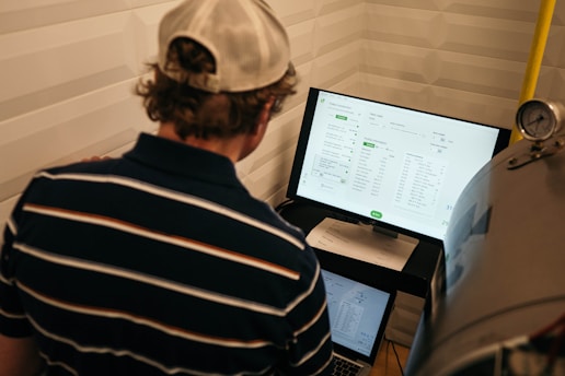 A person with a striped shirt and a cap is standing in front of a computer screen displaying data and analytics. Another laptop is open on a desk below the monitor, and a large metallic tank with a pressure gauge is nearby.