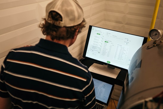 A person with a striped shirt and a cap is standing in front of a computer screen displaying data and analytics. Another laptop is open on a desk below the monitor, and a large metallic tank with a pressure gauge is nearby.