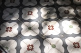 A pattern of decorative floor tiles with a floral motif, featuring white flower-like shapes centered with red and green elements. The tiles consist of repeated designs that create a geometric symmetry, and shadows cast across the surface add depth.