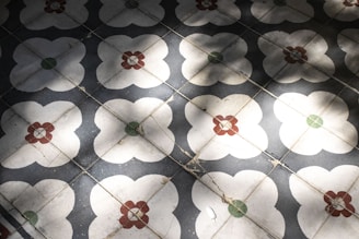 A pattern of decorative floor tiles with a floral motif, featuring white flower-like shapes centered with red and green elements. The tiles consist of repeated designs that create a geometric symmetry, and shadows cast across the surface add depth.
