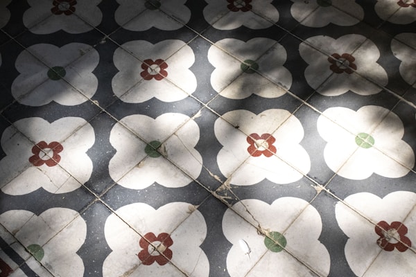 A pattern of decorative floor tiles with a floral motif, featuring white flower-like shapes centered with red and green elements. The tiles consist of repeated designs that create a geometric symmetry, and shadows cast across the surface add depth.