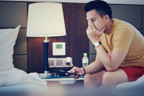 A person is seated on a bed in a modern room, looking at a smartphone. The individual appears deep in thought with one hand covering part of their face. Beside them, a lamp is lit on a nightstand. A water bottle and some electronic switches are visible in the background.