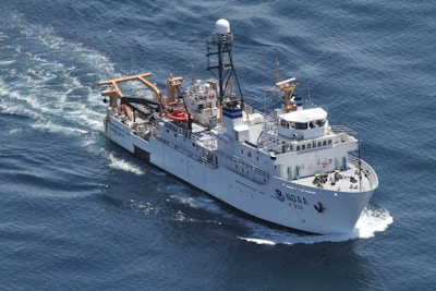A large research vessel navigates through the ocean, leaving a trail of white foam in its wake. The ship is equipped with various antennas and cranes, highlighting its function for scientific exploration. The vessel's name and identification number are visible on its side.