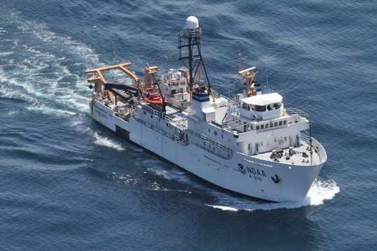 A large research vessel navigates through the ocean, leaving a trail of white foam in its wake. The ship is equipped with various antennas and cranes, highlighting its function for scientific exploration. The vessel's name and identification number are visible on its side.