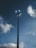 A tall streetlight with multiple fixtures extends into a partly cloudy blue sky. The light fixtures are evenly spaced around the top of the pole, and some are illuminated, creating a bright contrast against the sky.