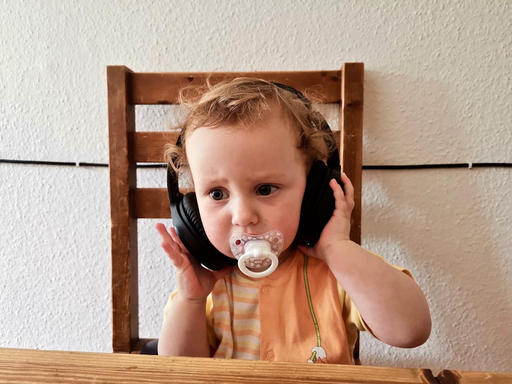 child with white pacifier using headphones while sitting on brown chair near table