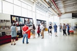 A group of people is gathered in a spacious, well-lit gallery or exhibition hall. They are observing artwork displayed on the walls, which include large paintings and smaller framed pieces. The environment features high ceilings and industrial-style elements, such as exposed pipes and large windows.