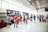 A group of people is gathered in a spacious, well-lit gallery or exhibition hall. They are observing artwork displayed on the walls, which include large paintings and smaller framed pieces. The environment features high ceilings and industrial-style elements, such as exposed pipes and large windows.