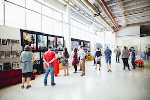 A group of people is gathered in a spacious, well-lit gallery or exhibition hall. They are observing artwork displayed on the walls, which include large paintings and smaller framed pieces. The environment features high ceilings and industrial-style elements, such as exposed pipes and large windows.