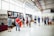 A group of people is gathered in a spacious, well-lit gallery or exhibition hall. They are observing artwork displayed on the walls, which include large paintings and smaller framed pieces. The environment features high ceilings and industrial-style elements, such as exposed pipes and large windows.